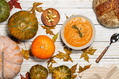 Pumpkin soup with rosemary twig aside chestnuts, pumpkins, bread and napkin on a white vintage wood background with autumn leaves. Flat Lay.