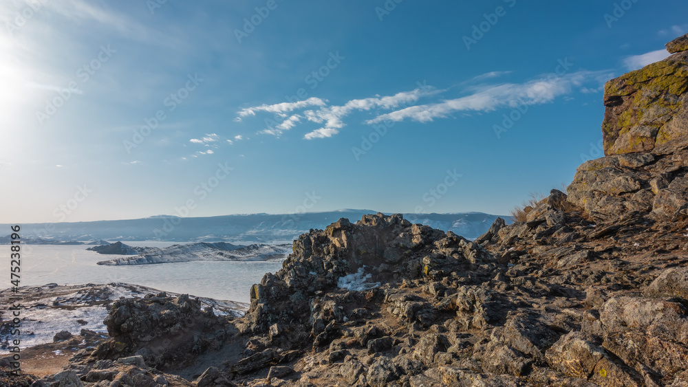 Winter Siberian landscape. In the foreground is a picturesque granite rock with cracked slopes. In the distance - a frozen lake, a mountain range. Blue sky with clouds. Baikal