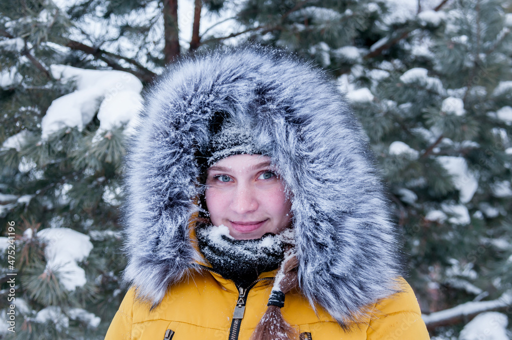 Fototapeta premium Portrait of a girl in a yellow down jacket with a fur hood on the background of a winter forest with fir trees