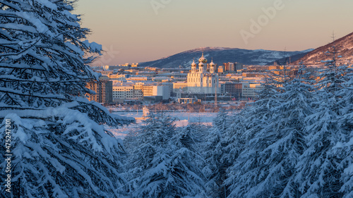 Amazing winter landscape. View from the snow-covered forest to the northern city and mountains. Snow on the branches of larch trees. Beautiful cathedral at sunrise. Cold snowy weather. Magadan, Russia photo