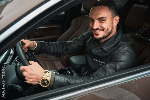 Portrait of handsome young man smiling while driving modern car. Driver wearing casual clothes and gold watch. © anatoliy_gleb