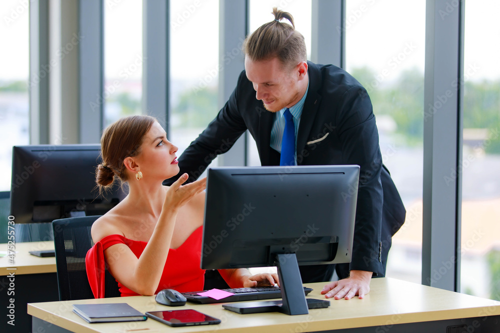 Caucasian beautiful sexy young female with braided bun hair in red luxury evening dress sitting at working desk with computer tablet and notebook asking male manager in formal suit in company office Caucasian beautiful sexy young female with braided bun hair in red luxury evening dress sitting at working desk with computer tablet and notebook asking male manager in formal suit in company office