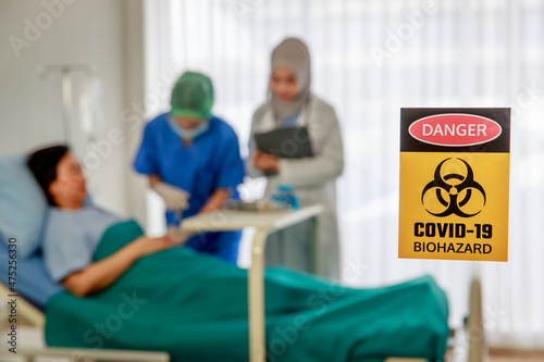 Closeup shot Danger Biohazard Covid-19 yellow hazard sign symbol banner in front blurred background of muslim Islam Arab doctor visit patient while nurse inject saline solution in hospital ward room