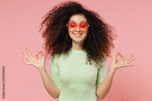 Young curly latin woman 20s wears mint t-shirt sunglasses hold spreading hands in yoga om aum gesture relaxing meditate try to calm down isolated on plain pastel light pink background studio portrait.