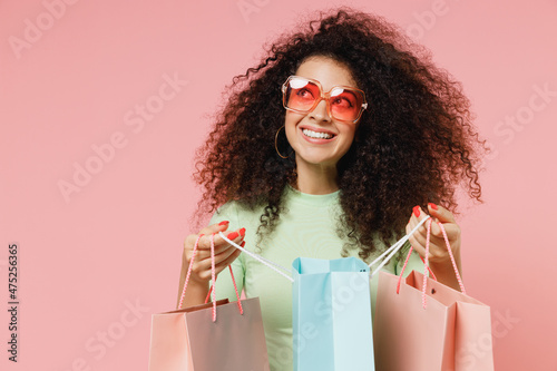 Dreamful fun young curly latin woman 20s wears mint t-shirt sunglasses looking aside holding package bags with purchases after shopping isolated on plain pastel light pink background studio portrait.