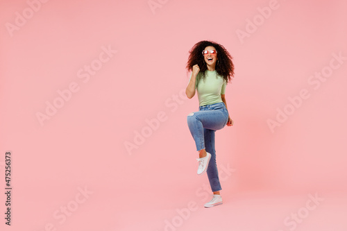 Full size body length young curly latin woman 20s wears casual clothes sunglasses doing winner gesture celebrate clenching fists say yes isolated on plain pastel light pink background studio portrait.