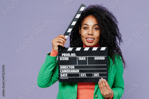 Charming happy charismatic smiling vivid young black curly woman 20s wears green shirt holding classic black film making clapperboard isolated on plain pastel light violet background studio portrait.