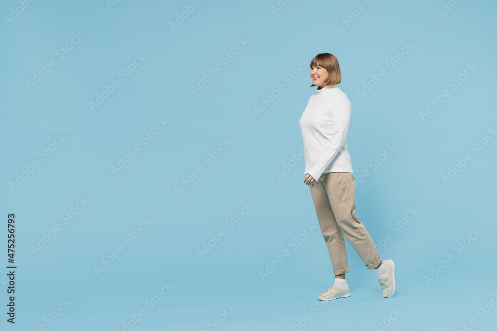 Full body side view elderly smiling cheerful fun caucasian woman 50s wear white knitted sweater walking going stroll isolated on plain blue color background studio portrait. People lifestyle concept.