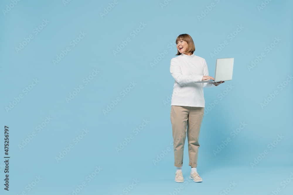 Full body elderly smiling woman 50s wearing white knitted sweater hold use work on laptop pc computer look aside on copy space area isolated on plain blue color background. People lifestyle concept.