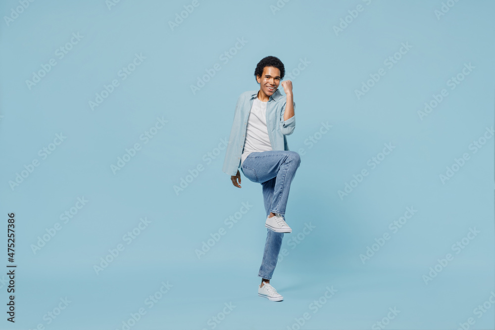 Full size body length vivid young black curly man 20s years old wears white shirt doing winner gesture celebrate clenching fists say yes isolated on plain pastel light blue background studio portrait.