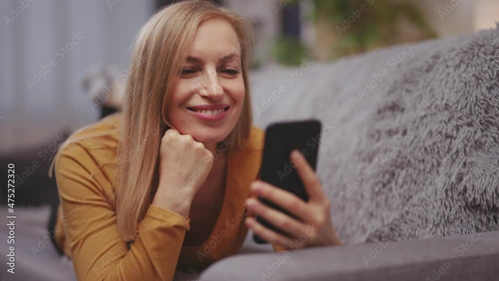 Caucasian middle aged woman lying on cozy grey sofa with modern smartphone on hands. Pleasant blonde using gadget for browsing internet at home. Caucasian middle aged woman lying on cozy grey sofa with modern smartphone on hands. Pleasant blonde using gadget for browsing internet at home.