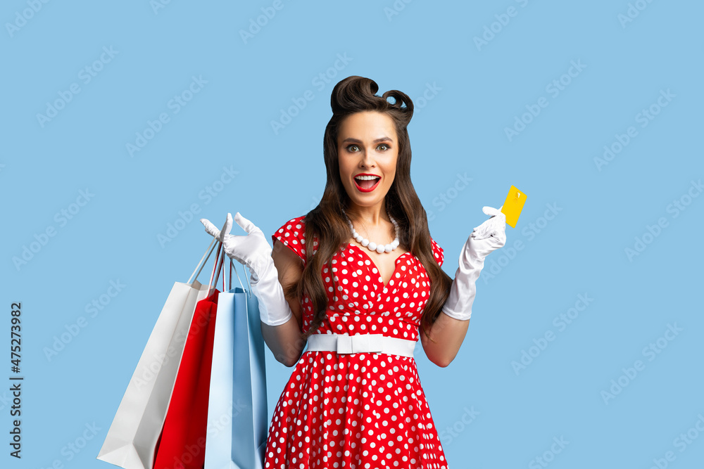 Attractive young pinup woman in retro style dress holding gift bags and credit card over blue studio background Attractive young pinup woman in retro style dress holding gift bags and credit card over blue studio background