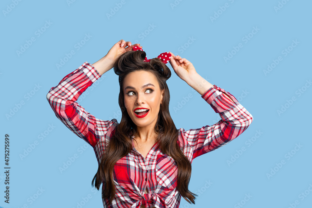 Attractive pinup lady in retro outfit tying bow on her head, styling her hair on blue studio background Attractive pinup lady in retro outfit tying bow on her head, styling her hair on blue studio background