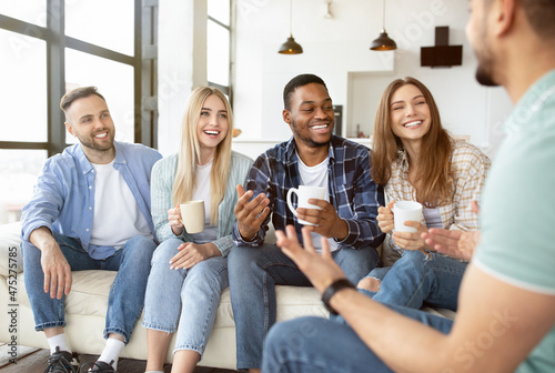 Diverse millennial friends talking and laughing together, drinking tea or coffee at home