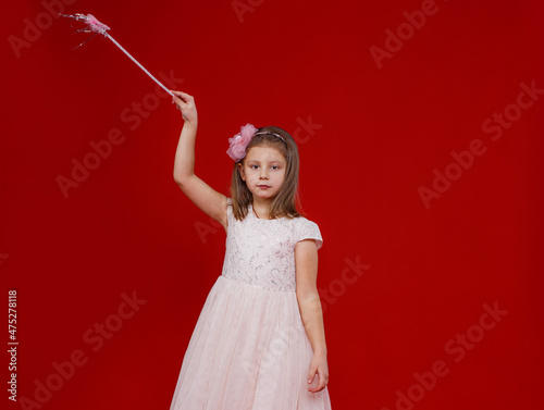 a beautiful little girl in a fairy dress with a magic wand in her hands stands on a red isolated background.
