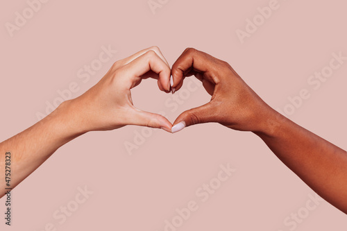 Close up of black and white female hands in heart shape  interracial friendship. Multiracial hands over beige background at studio. Peace and love concept.