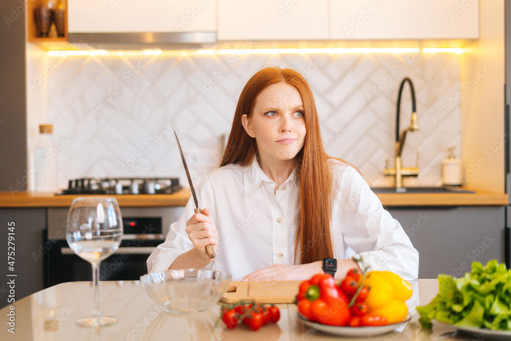 Portrait of attractive redhead young woman with dissatisfied eyes holding big knife sitting at table with cutting board, looking away in light kitchen room with modern interior, close-up.