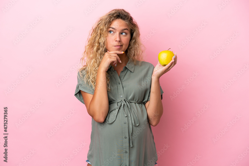 Girl with curly hair isolated on pink background pregnant and holding an apple while thinking Girl with curly hair isolated on pink background pregnant and holding an apple while thinking