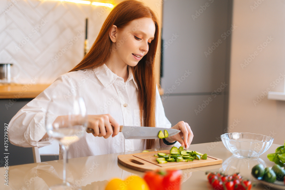 Close-up side view of attractive young redhead woman cutting fresh cucumber cooking food salad sitting at table in kitchen room. Happy female cooking vegetarian dieting salad full of vitamins.