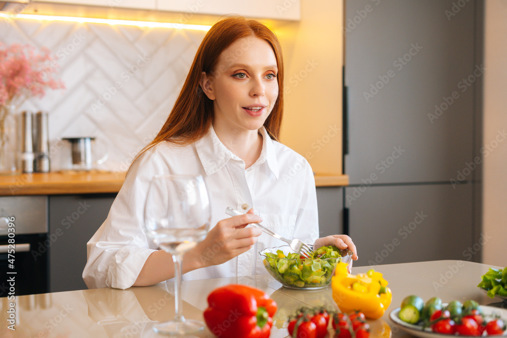 Pretty happy young redhead woman eating fresh vegetarian salad enjoying fresh tasty vegetables sitting at table in kitchen room with modern light interior. Concept of healthy lifestyle and eating.