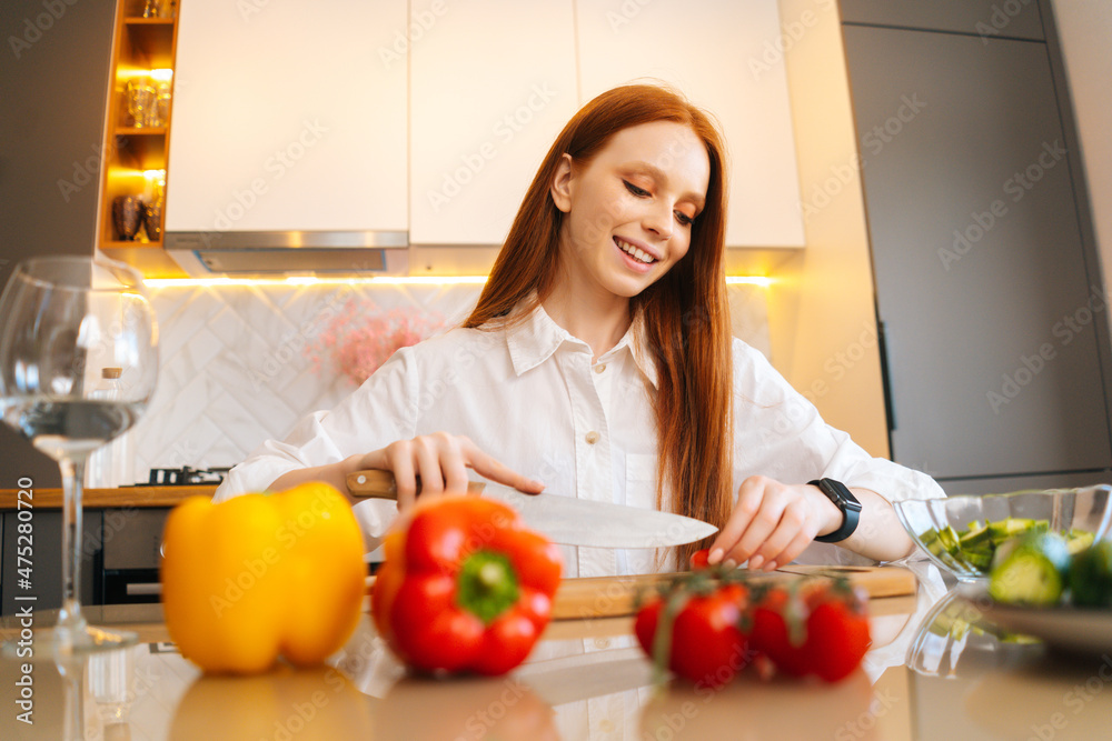 Portrait of cheerful young redhead woman cutting fresh tomato preparing food salad sitting at table in modern kitchen room. Happy female cooking vegetarian dieting salad full of vitamins. Portrait of cheerful young redhead woman cutting fresh tomato preparing food salad sitting at table in modern kitchen room. Happy female cooking vegetarian dieting salad full of vitamins.