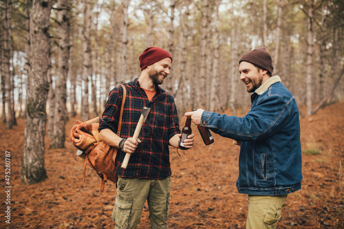 Two millennial man hiking and drinking beer in a forest, laughing and having fun. © polinaloves