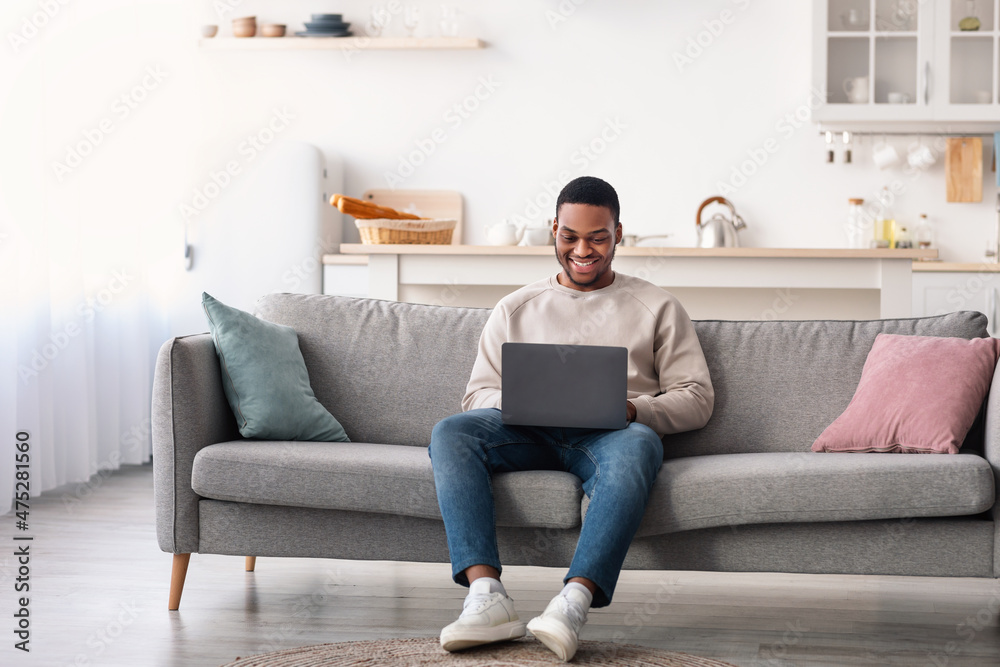 Smiling black guy using laptop, sitting on couch