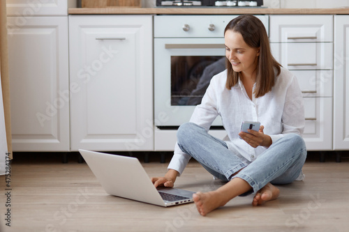 Portrait of smiling woman using laptop computer, freelancer working online, female wearing white t shirt and jeans sitting on floor in kitchen, holding mobile phone in hands.