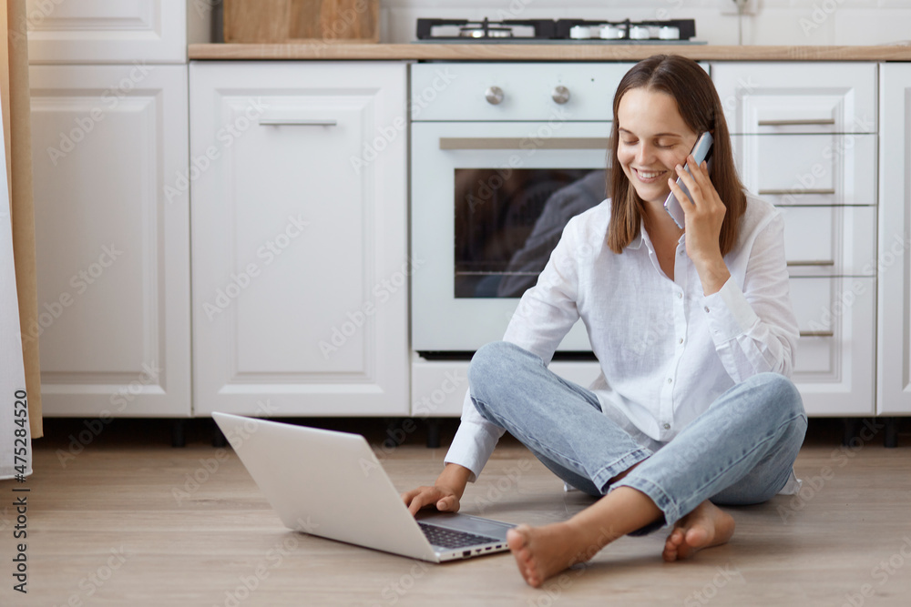 Image of winsome young adult Caucasian woman wearing white t shirt and jeans sitting on floor in kitchen, talking phone, working on notebook, blogger writing posts for social networks. Image of winsome young adult Caucasian woman wearing white t shirt and jeans sitting on floor in kitchen, talking phone, working on notebook, blogger writing posts for social networks.