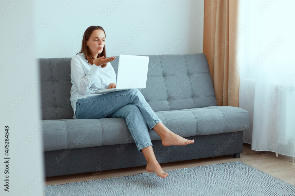 Indoor shot of happy romantic dark haired woman sitting on sofa with laptop, wearing white shirt and jeans, looking at notebook screen while having video call with husband, sending air kissing. Indoor shot of happy romantic dark haired woman sitting on sofa with laptop, wearing white shirt and jeans, looking at notebook screen while having video call with husband, sending air kissing.