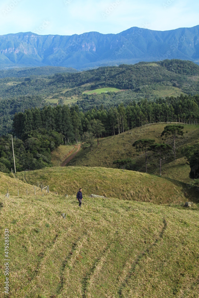 turista não identificado caminhando na grama com montanhas ao fundo em Anitápolis, Santa Catarina