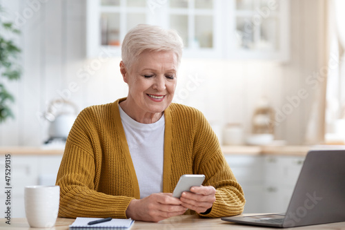 Smiling mature lady sitting at table with laptop, using smartphone photo