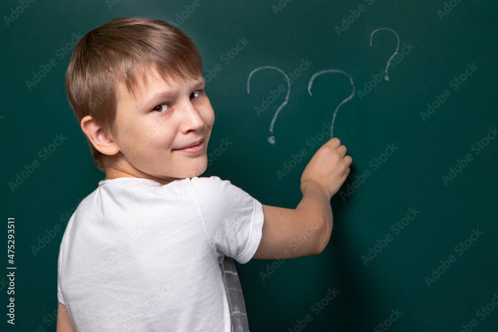 A boy of Caucasian appearance writes a question mark on the chalkboard. Looking into the camera. Smile. Easy solution to the problem.