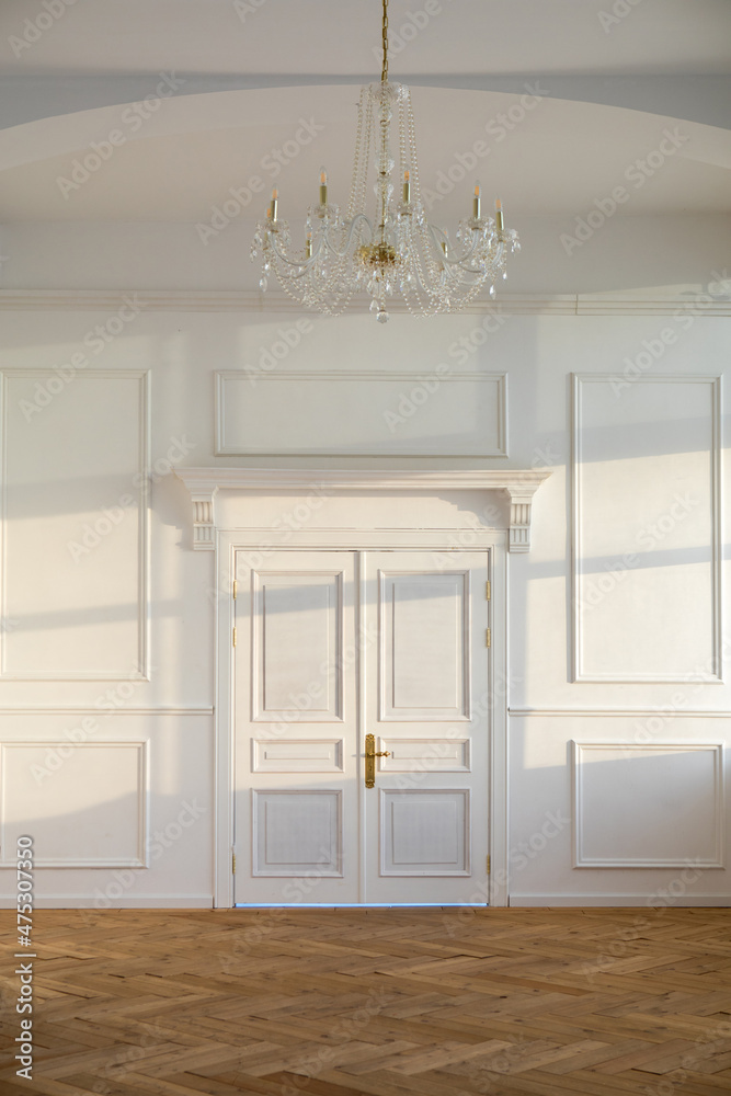 White closed doors in light spacious empty room with classic design and chandelier hanging on ceiling near wall with sunlight