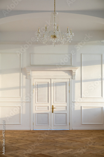 White closed doors in light spacious empty room with classic design and chandelier hanging on ceiling near wall with sunlight