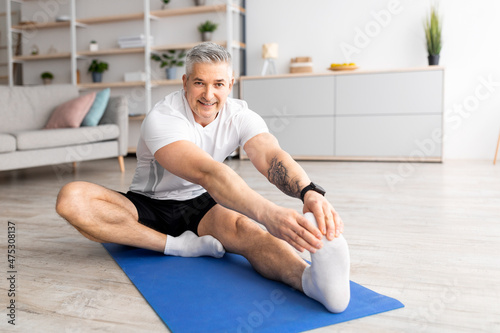Sport at home during quarantine. Senior man doing stretch exercises for legs sitting on mat in living room