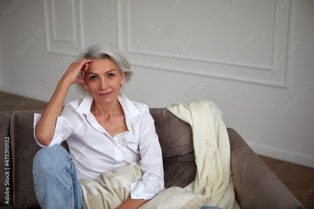 Smiling stylish mature female with gray hair sitting on sofa and leaning on hand while chilling in living room and looking at camera Smiling stylish mature female with gray hair sitting on sofa and leaning on hand while chilling in living room and looking at camera