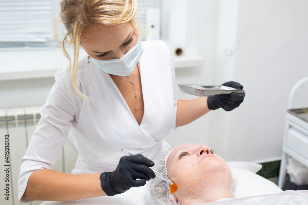 Portrait of a young dermatologist in a medical mask and gloves applying a special gel with a brush to the face of a lying female client before the procedure of non-surgical ultrasound SMAS lifting Portrait of a young dermatologist in a medical mask and gloves applying a special gel with a brush to the face of a lying female client before the procedure of non-surgical ultrasound SMAS lifting