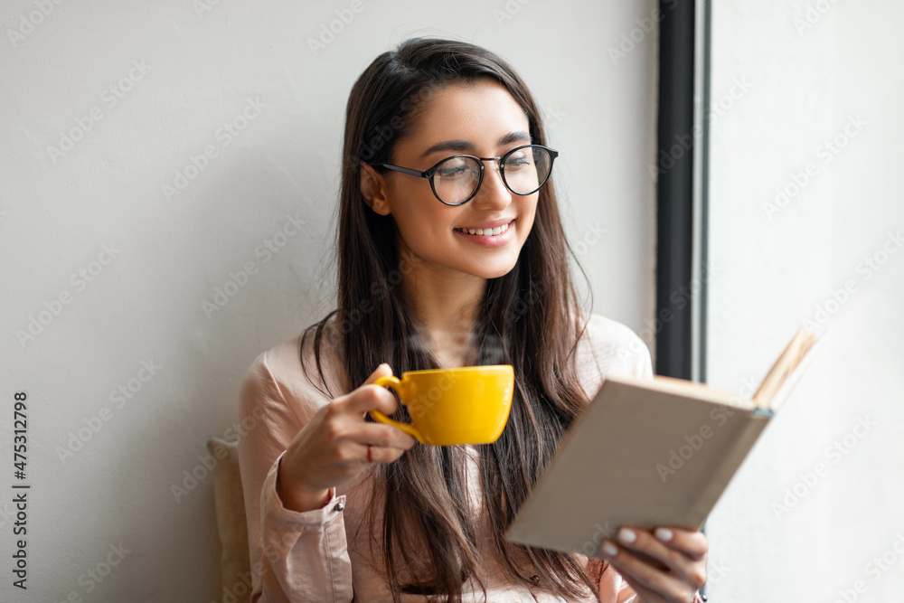 Leisure lifestyle concept. Excited arab lady with mug of coffee reading book while resting in cafe near window Leisure lifestyle concept. Excited arab lady with mug of coffee reading book while resting in cafe near window