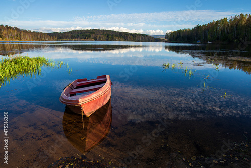 A red old wooden boat anchored near the shore, in a secluded harbor. Landscape photography of a beautiful blue lake and an anchored red boat.