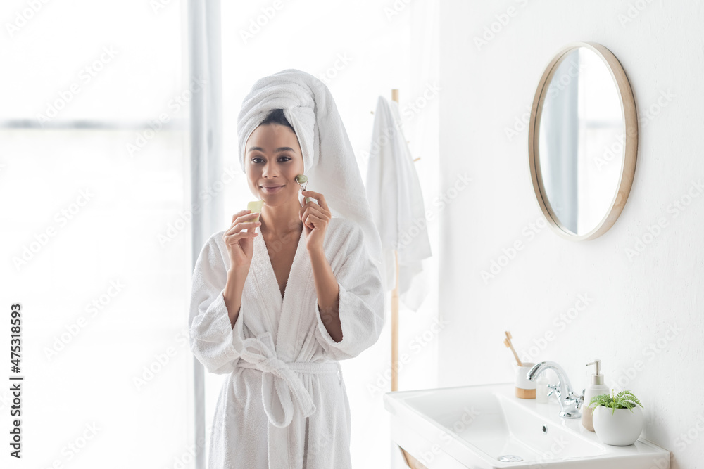 happy african american woman in bathrobe massaging face with jade roller and face scraper happy african american woman in bathrobe massaging face with jade roller and face scraper