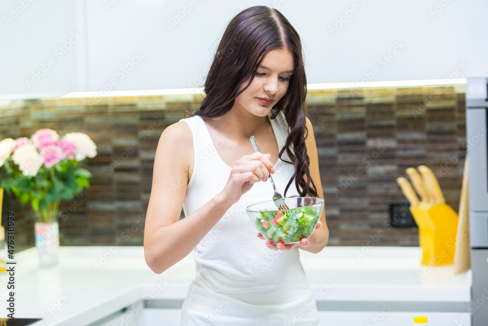 Portrait of a cute brunette girl standing in a white top in the kitchen and eating a healthy vegetable vitamin salad from a glass bowl for breakfast Portrait of a cute brunette girl standing in a white top in the kitchen and eating a healthy vegetable vitamin salad from a glass bowl for breakfast
