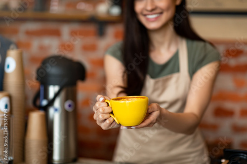 Unrecognizable female barista welcoming guests at coffee house counter, offering cup of hot beverage, closeup, crop