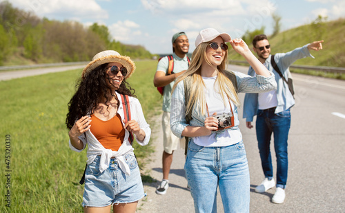 Group of positive young friends walking along highway, making photos, having fun together outdoors