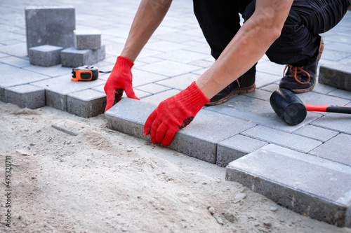 The master in yellow gloves lays paving stones in layers. Garden brick pathway paving by professional paver worker. Laying gray concrete paving slabs in house courtyard on sand foundation base.