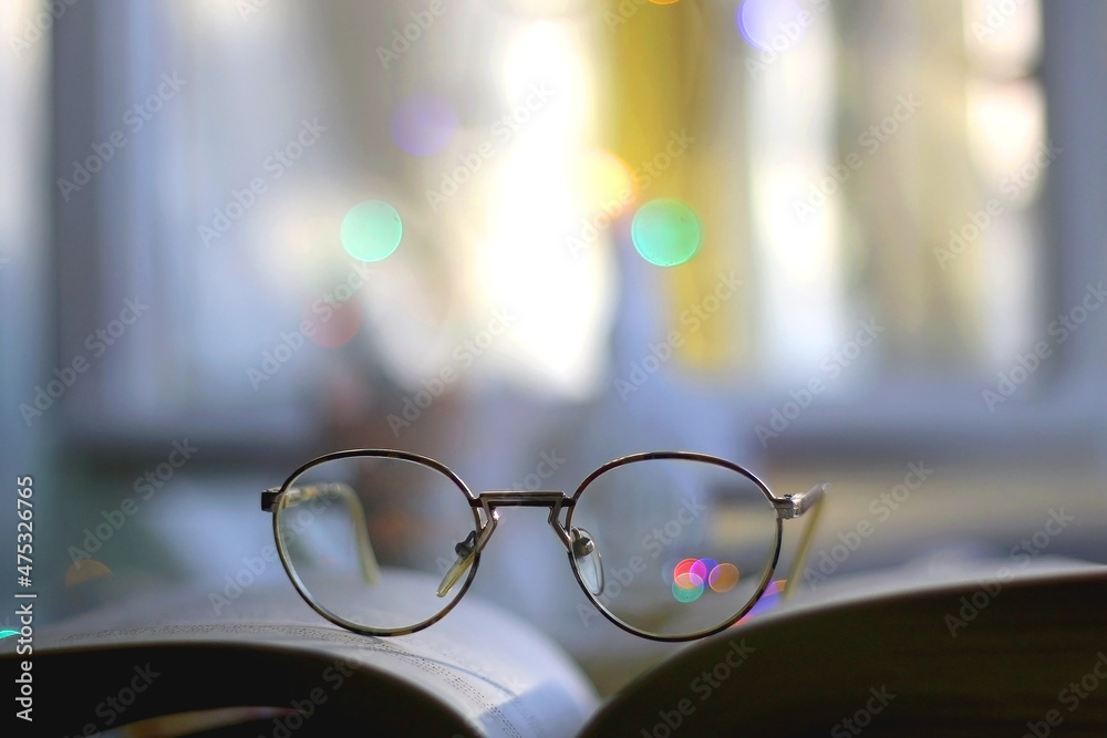 Open book and reading glasses with candles and colorful bokeh lights in the background. Selective focus. Open book and reading glasses with candles and colorful bokeh lights in the background. Selective focus.