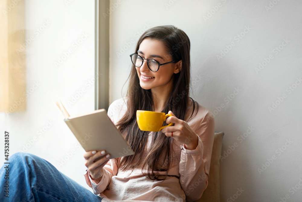 Relaxed arab woman reading favorite book and drinking coffee, resting in cafe near window, free space. Relaxed arab woman reading favorite book and drinking coffee, resting in cafe near window, free space.
