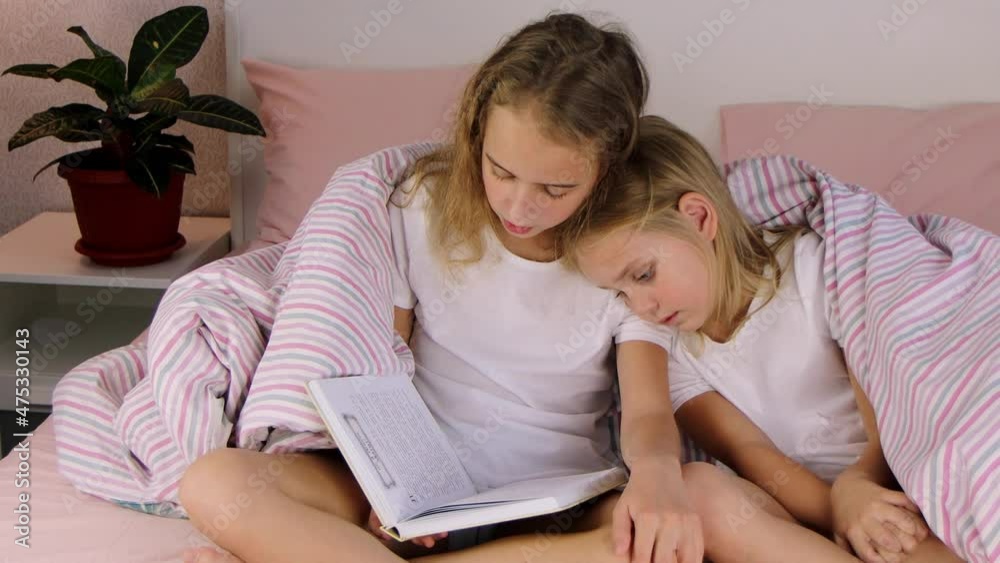 two cute little girls sisters are sitting on the bed covered with a warm blanket and reading a children's book, one girl put her head on the shoulder of the other, family values and traditions two cute little girls sisters are sitting on the bed covered with a warm blanket and reading a children's book, one girl put her head on the shoulder of the other, family values and traditions