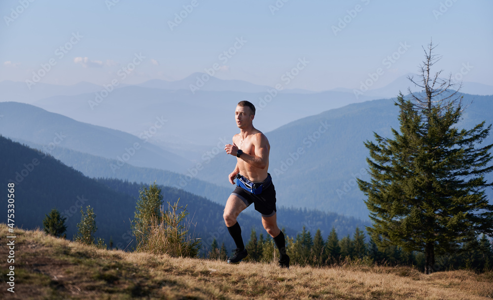 Young man running up grassy hill. Muscular male trail runner wearing black sports shorts while jogging outdoors in mountains. Concept of sport, active leisure and nature.