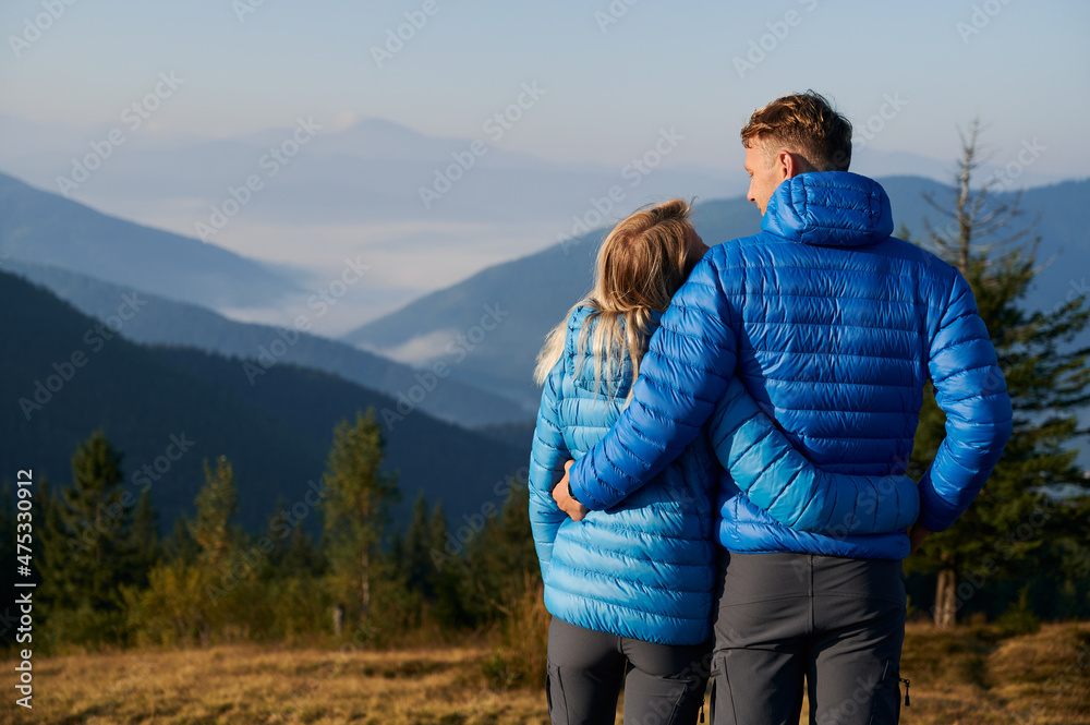 Rear view of lovers hugging and cute looking at each other against the backdrop of beautiful mountain beskids, while walking outdoors in sunny early autumn weather.